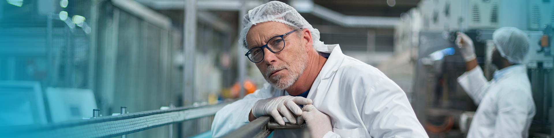Ein Mann mit weißer Schutzkleidung, Haarnetz und Handschuhen wirft einen prüfenden Blick auf die Produktionsanlage.