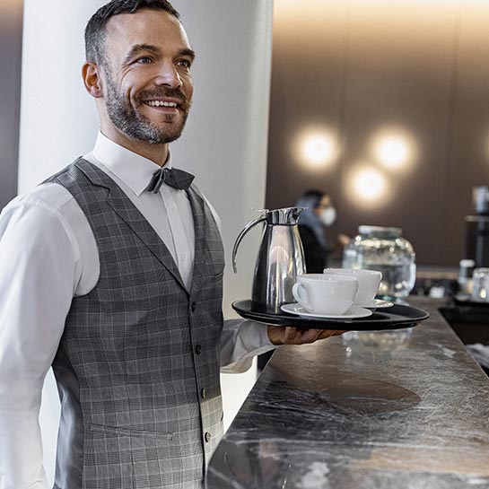Gastromitarbeiter in weißem Hemd, grauer Weste und passender Fliege steht an einem Tresen und ält ein Tablett mit einem Kaffee-Service in der Hand.