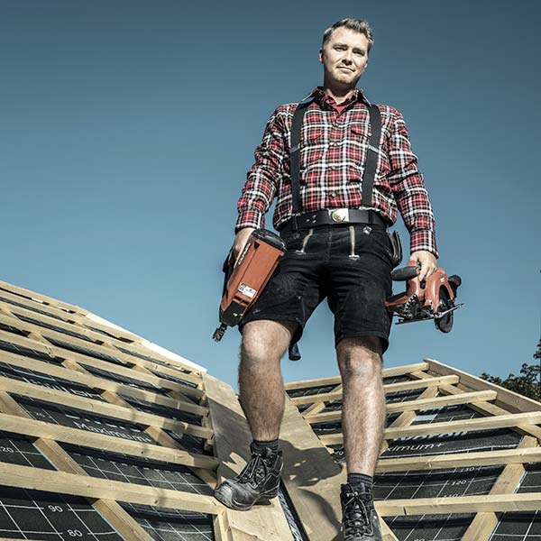 Dachdecker in kurzer Hose und kariertem Hemd steht auf einem Dachstuhl und hält Werkzeug in der Hand.