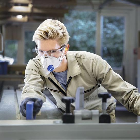 Eine blonde Frau mit kurzen Haaren richtet ein Element an einer Maschine aus und trägt dabei eine durchsichtige Schutzbrille sowie eine Atemschutzmaske von Dräger.