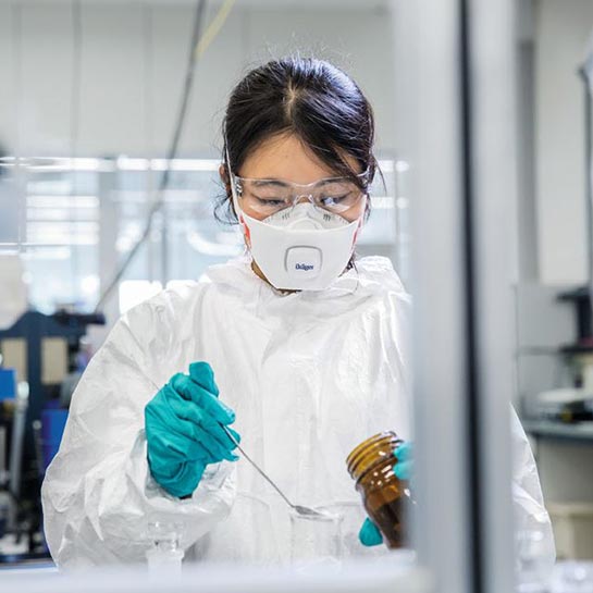 Eine Frau mit dunklen Haaren steht in einem Labor, mischt etwas zusammen und trägt dabei eine Schutzbrille sowie einen Atemschutz.