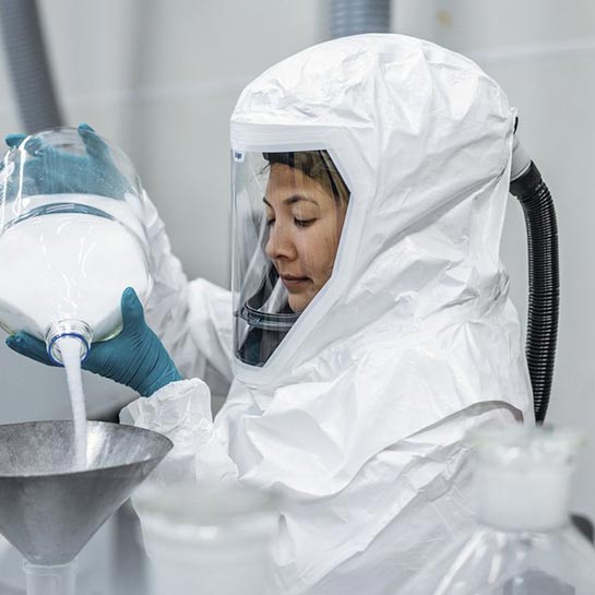 Frau trägt einen Ganzkörperschutzanzug mit Luftsystem und füllt ein weißes Pulver in einen Edelstrahl-Trichter. 