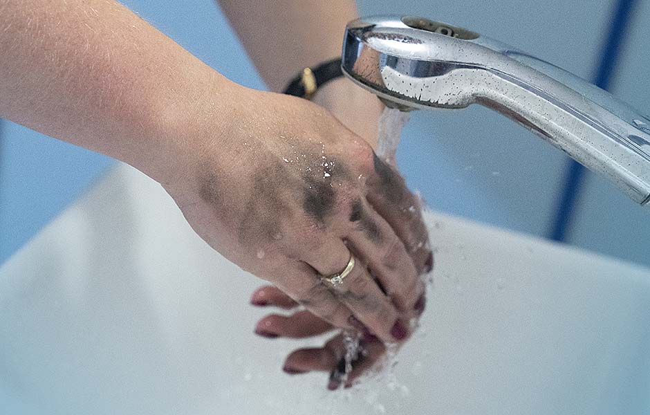 Zwei dreckige Hände mit roten Fingernägeln, goldenem Verlobungsring und schwarzer Uhr werden unter fließendem Wasser an einem Waschbecken gewaschen. 